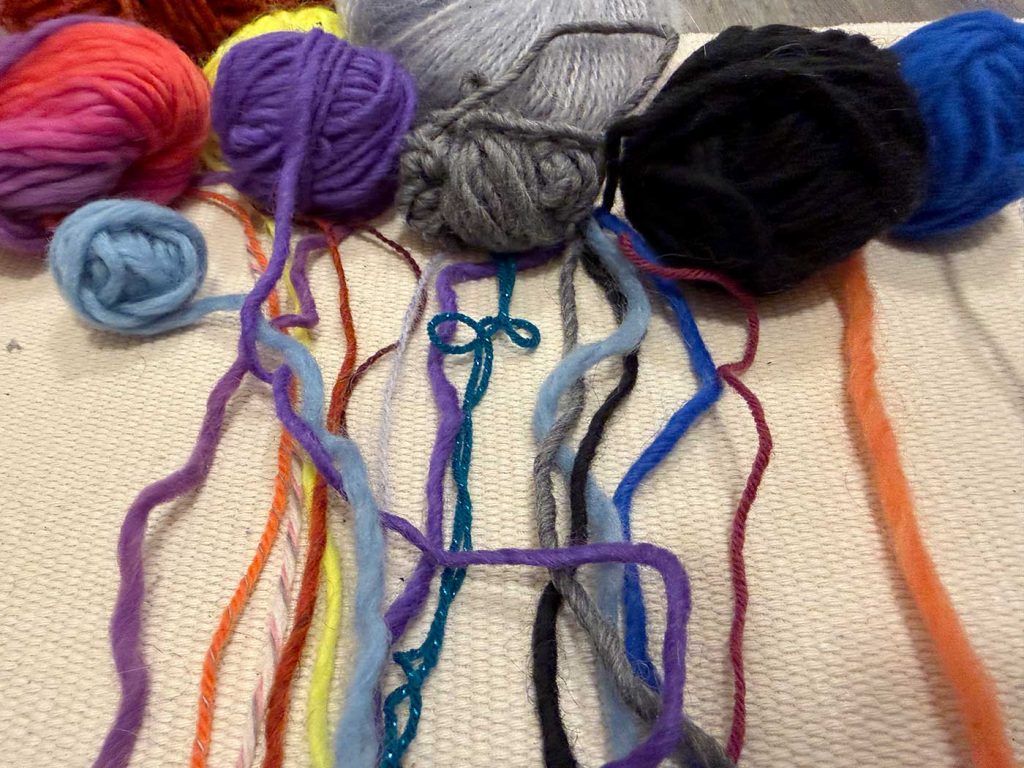Nystan med garn i olika färger och tjocklekar. Från nystanen har lite garn dragits ut, och trådarna ligger lite ojämnt fördelat på ett ljust underlag.
