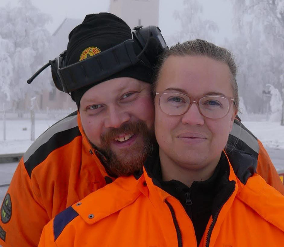 En porträttbild av en kvinna och en man som står tätt intill varandra. Bägge ser glada ut och har varselkläder på sig.