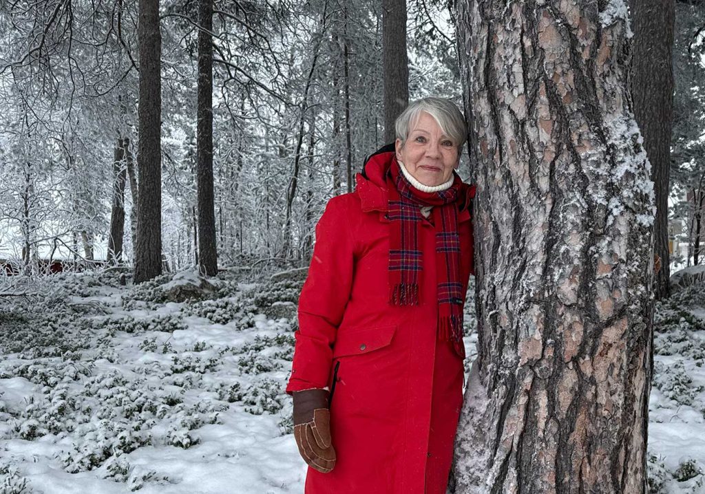 En leende kvinna lutar sig mot en tallstam med grov, skrovlig bark. Hennes blick är riktad rakt mot fotografen, och i den röda jackan sticker hon tydligt ut mot bakgrunden, med delvis snötäckt skogsmark och rimfrost på grenarna. 