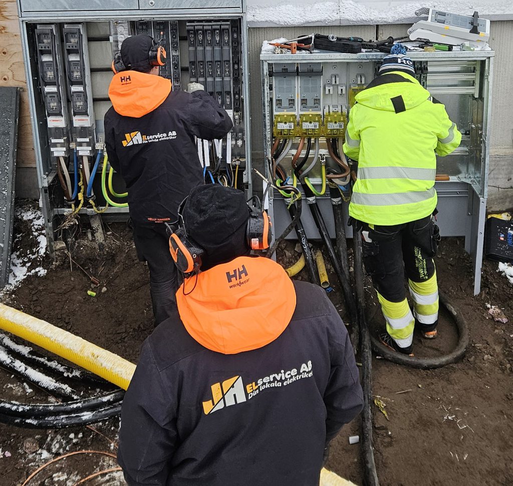 Bild som visar tre män bakifrån, utomhus. De jobbar i ett elskåp med många kablar och kopplingar. Två av männen har jackor med logga för JA Elservice AB. 