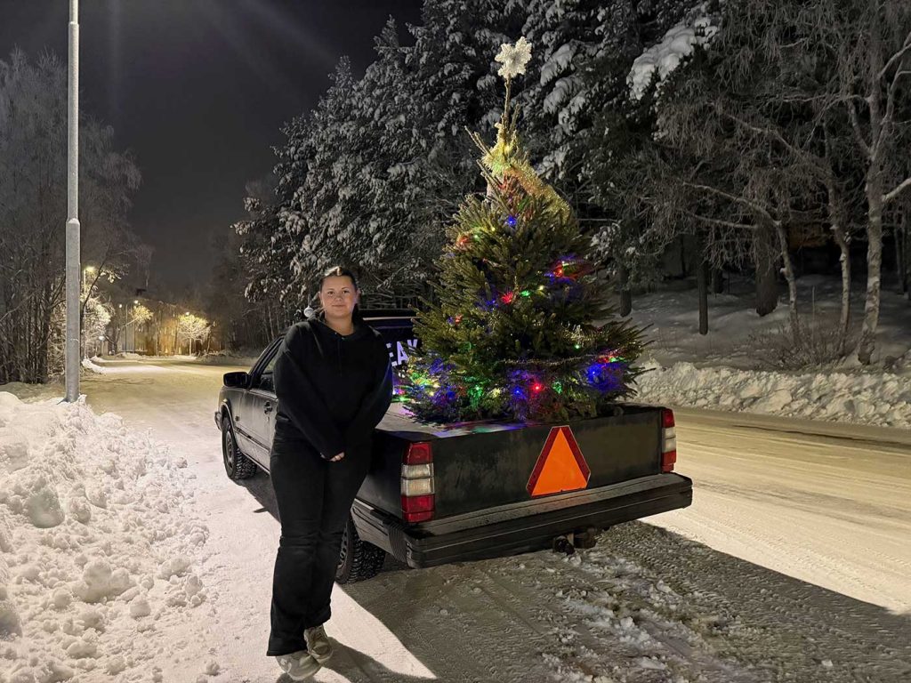 En svartklädd ung tjej bredvid en svart EPA med en julgran på flaket. Julgranen är klädd med en färgglad ljusslinga som lyser i mörkret.