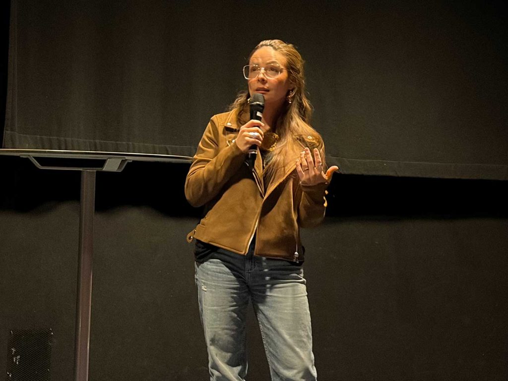 En kvinna med mikrofon i hand, nära munnen. Hon ser tankfull ut, har blicken fäst på något långt borta och gestikulerar med den hand som inte håller i mikrofonen. 