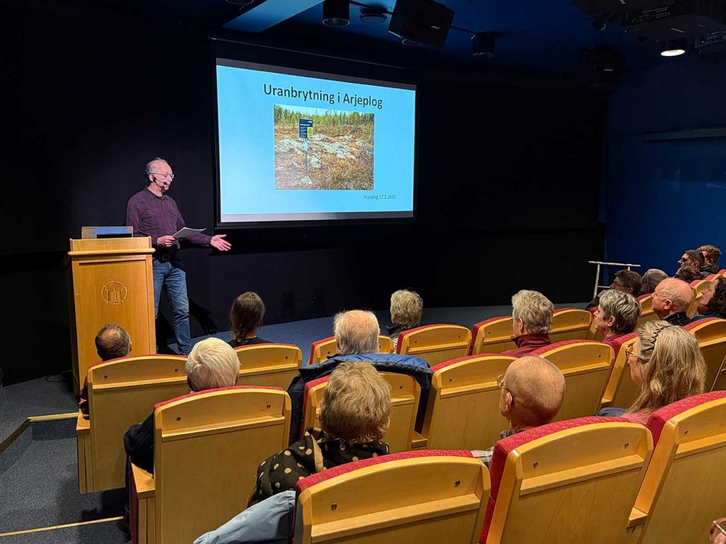 I förgrunden syns publik i en föreläsningssal bakifrån. På scenen står en man i rutig skjorta och jeans vid en talarstol. Bakom honom på en bildskärm syns ett foto på en varningsskylt som LKAB satt upp i Pleutajokk. Ovanför bilden står texten Uranbrytning i Arjeplog.