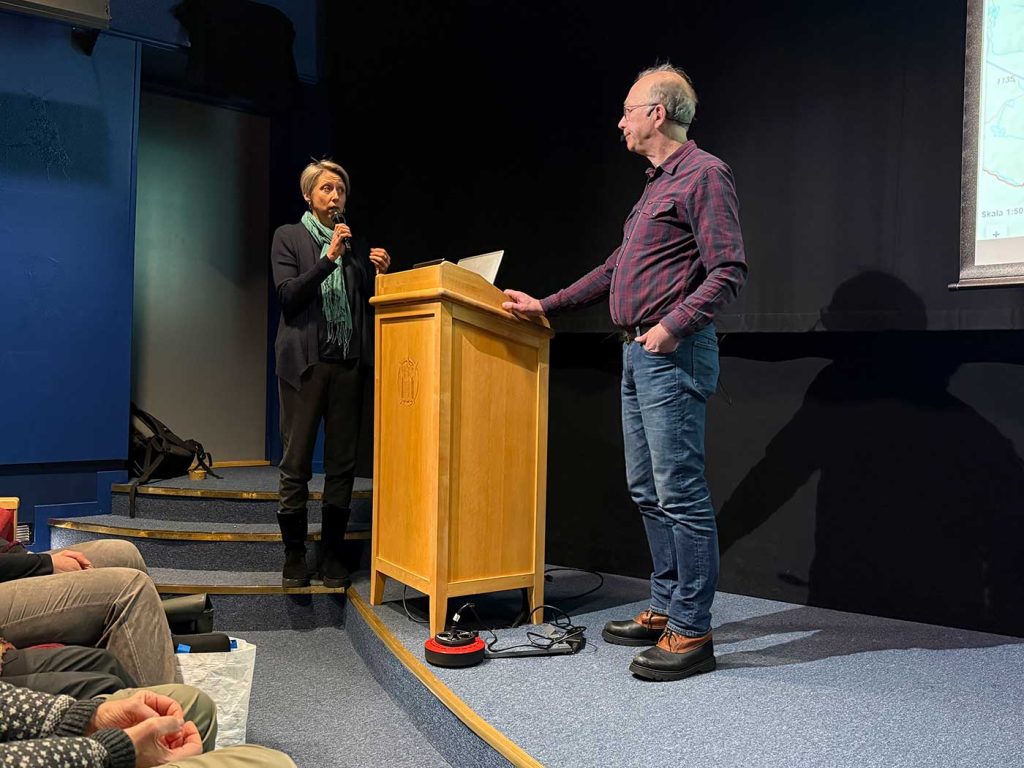 En talarstol mitt i bilden. TIll vänster om den står en kvinna och pratar i en mikrofon. Hon har blicken fäst på en man som står till höger om talarstolen. 