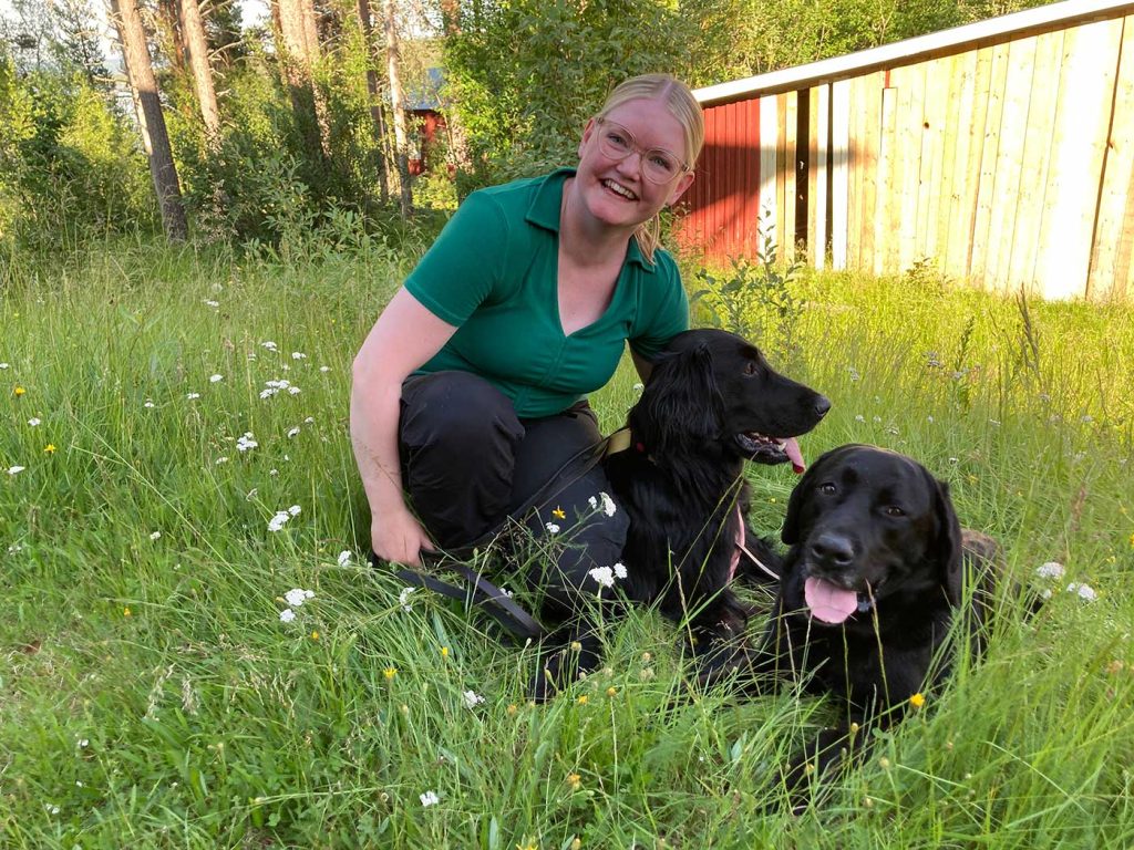 Åsa står på huk i grönt gräs, med hundarna Nemo och Herman liggandes bredvid. Åsa tittar in i kameran och skrattar. Hundarna har öppna munnar och tungorna hänger ut. 