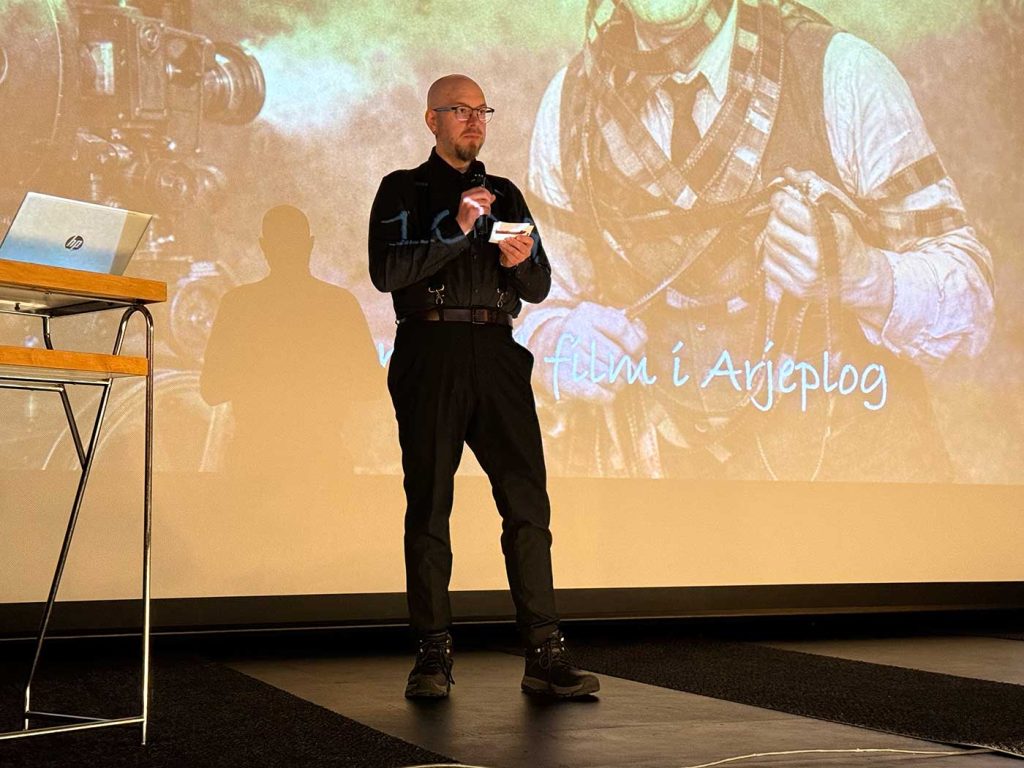 En man står på en scen framför en bioduk. På duken syns en illustration som ser ut som ett gammalt foto, av en man som trasslat in sig i filmremsor. Mannen tittar rakt framåt, håller en mikrofon i en handen, nära munnen, och små anteckningspapper i andra handen. 