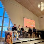 Vybild inifrån en konferenssal. På scenen sitter sju personer på höga pallar. TIll vänster i bild syns ett stort fönster.