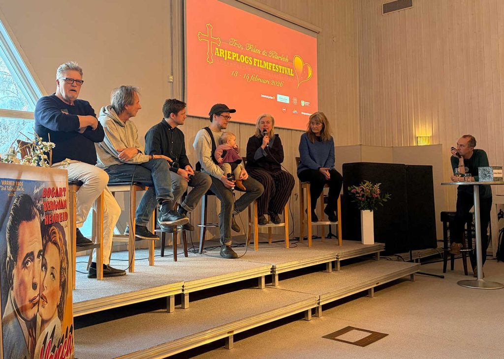 Sex personer sitter på höga pallar på en liten scen. En av dem, en kvinna, håller en mikrofon nära sin mun. Dom andra riktar sin uppmärksamhet mot henne. Bakom dem på väggen syns en bild med texten Tro, Film och Kärlek Arjeplogs Filmfestival 13-15 februari 2026. Längst till höger i bild, nedanför scenen, står en man lutad mot ett högt bord. Han håller en mikrofon i handen och tittar på kvinnan som pratar. 