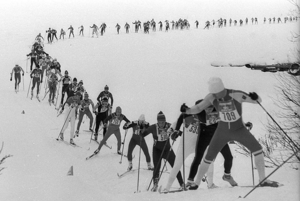 En lång rad med skidåkare kommer längs en sjö och åker uppför en backe, mot fotografen.