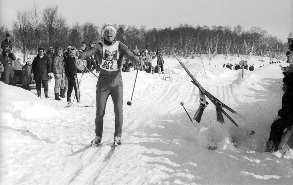 En svartvit gammal bild från en skidtävling. En skidåkare med öppen mun, som ser ut att ta i av alla krafter, är på väg mot fotografen. I spåret bredvid honom ser man skidor, ben och en stav på en annan skidåkare som precis kört omkull. Resten av kroppen är dold i virvlande snö. I bakgrunden syns publik.