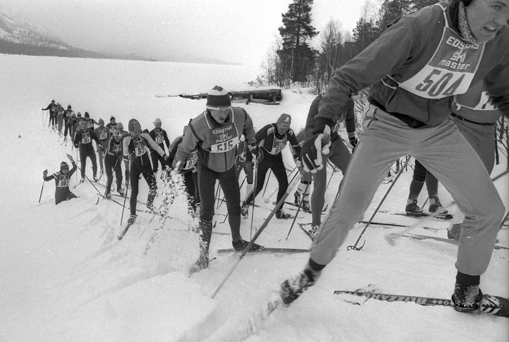 En grupp skidåkare stakar sig uppför en backe. Det är en svartvit gammal bild. Alla har nummerlappar på sig.