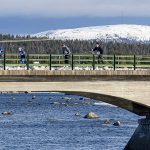 En bild tagen på avstånd. Skidåkare passerar en bro, och döljs delvis bakom räcket. under bron öppet vatten.