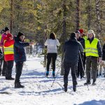 Bild på människor som står på båda sidor av ett skidspår i skog. I spåret syns två åkare på väg bort mot fotografen. En person håller fram en mugg mot en av skidåkarna, och till höger i bild syns ett bord med muggar, vattenflaskor och en vattendunk.