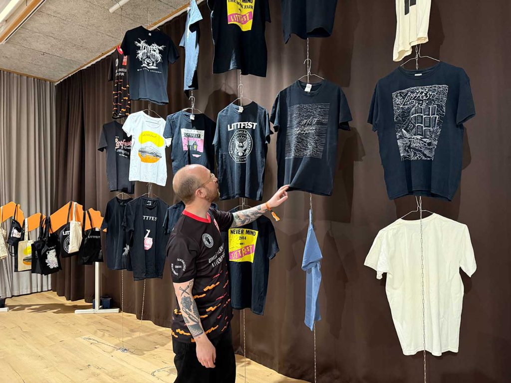 T-shirts med olika motiv hänger ner från taket. En man (Hugo Sundkvist) håller i och tittar på en av dem samtidigt som han säger något.