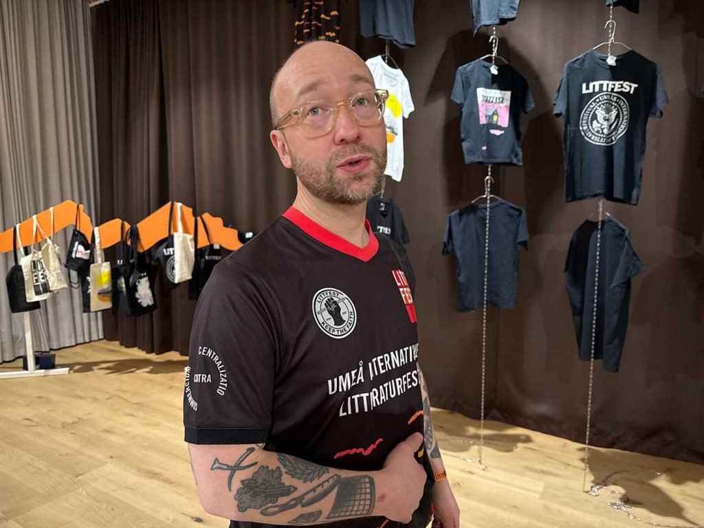 Bild av en man som står lite snett vänd mot fotografen, och ser ut att säga något. Bakom honom syns tygväskor och t-shirts som delar av en utställning med marknadsmaterial för Littfest.