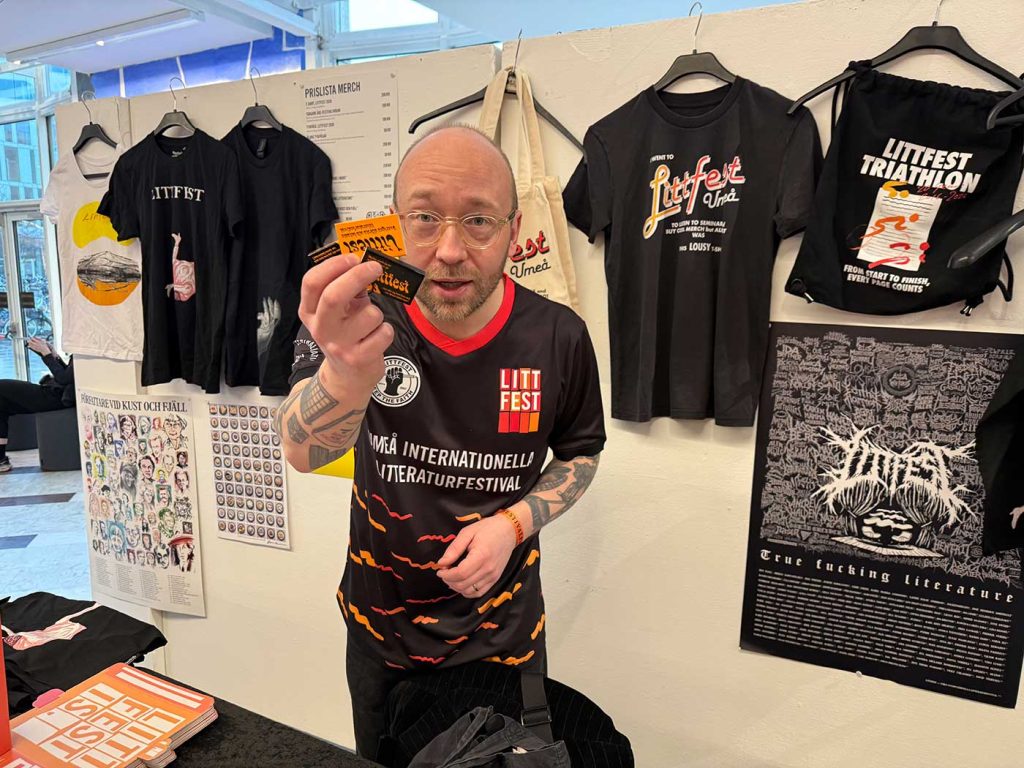 Ett försäljningsstånd med reklam för Littfest, bland annat t-shirts och tygväskor. Mitt i bilden står en man. Han skrattar och håller fram några tändsticksaskar med texten Littfest.
