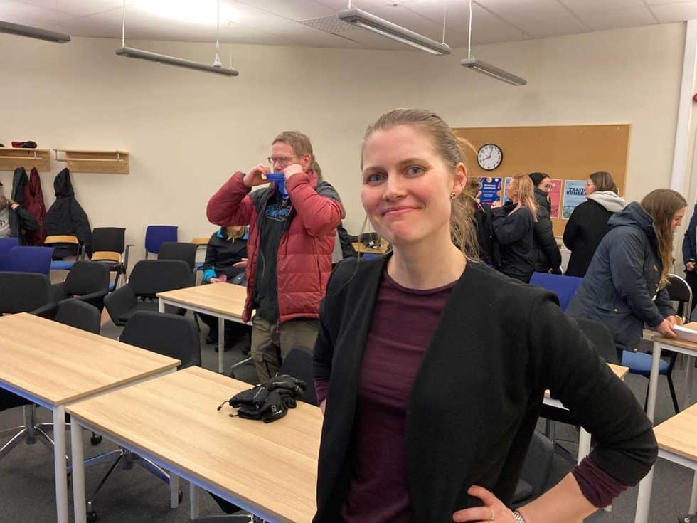 I förgrunden en bild på en kvinna, hon har händerna i sidorna, ler mot kameran och ser ganska bestämd, självsäker ut. Bilden är tagen i en möteslokal, och bakom kvinnan syns bord och människor som tar på sig ytterkläder.