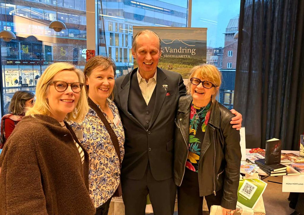 Tre kvinnor och en man står tätt intill varandra och tittar mot kameran. Alla är finklädda och ser väldigt glada ut. Genom fönsterglaset bakom dem syns en byggnad med många fönster. Det finns också en rollup bakom dem med reklam för Bok och vandring. Det man ser av rollupen visar ett par vandrare i fjällmiljö och orden ”Vandring” samt ”Jäggeluaktta”.