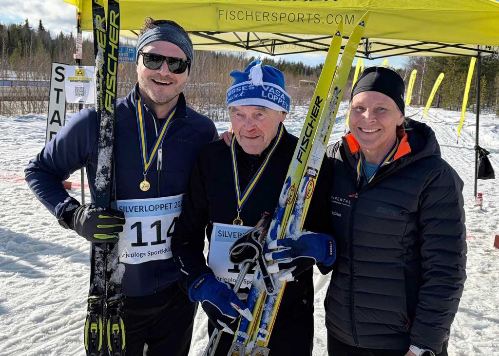 Tre glada människor, två män och en kvinna. De bägge männen håller i sina skidor, de har nummerlappar och medaljer på sig.