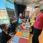 En översiktsbild över utställningsplatsen på Littfest i Umeå. I förgrunden ett bord med trycksaker, två kvinnor sitter bakom bordet och pratar glatt med en man som står på andra sidan av bordet. Det bortre bordet tillhör Bok och Vandring och är fyllt av böcker och reklammaterial. En kvinna lutar sig fram över bordet och pratar med LInda som håller Magasin Silvervägen i handen.