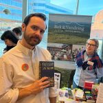 En leende man (författaren Carl Åkerlund) i vit skjorta. Han håller i sin bok Fädernesland. Bakom honom bokbord, en rollup med bilder från Bok och Vandring och en skrattande kvinna (Linda Buska)