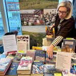 Ett bord fyllt med böcker och reklam. En kvinna står bredvid bordet och rättar till en broschyr som ligger snett. I bakgrunden en rollup med bilder från tidigare års Bok och Vandring.
