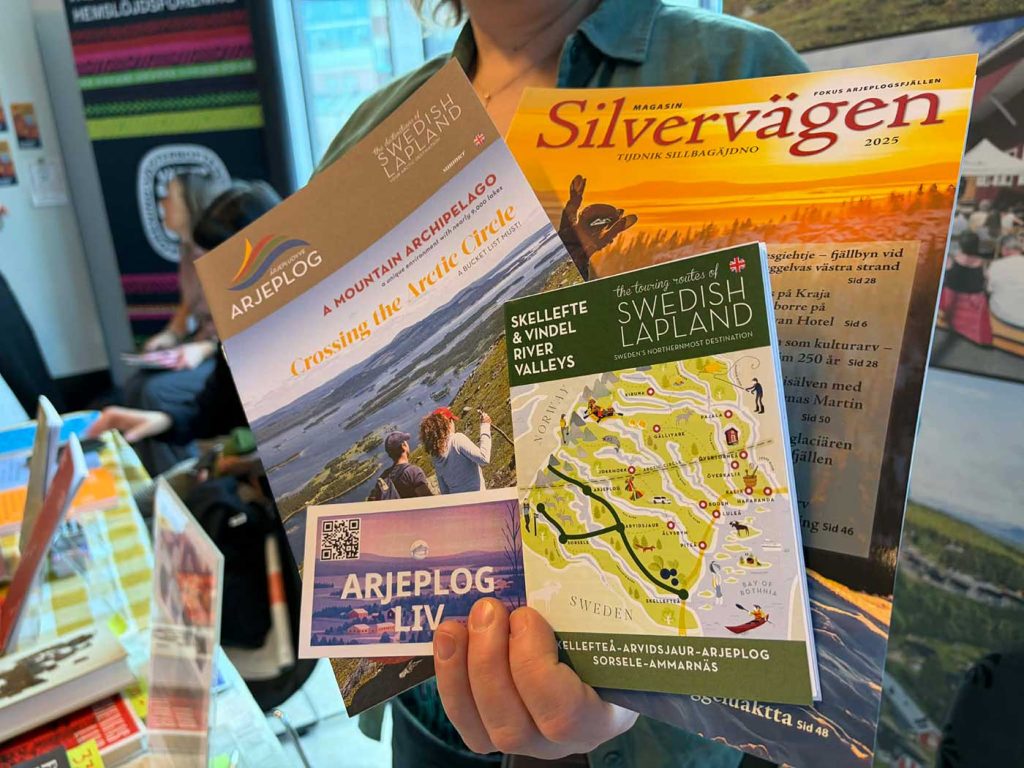 Närbild på reklam för Arjeplog. En hand håller upp Magasin Silvervägen, en broschyr om Arjeplog, ett häfte med en karta över Skellefteå och Vindelälvens dalgångar, samt ett litet visitkort med en QR-kod och texten Arjeplogliv.