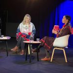Två kvinnor (Kathrine Nedrejord och Elin Anna Labba) sitter i varsin fåtölj på en scen. Elin Anna håller i Kathrines bok Sameproblemet och säger något som får Kathrine att skratta. På låga bord bredvid dem står glas och karaffer med vatten.