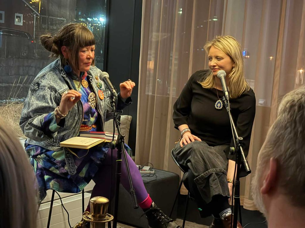 Två kvinnor sitter på varsin hög stol i ett hörn. Bakom den ena kvinnan finns ett fönster ut mot en mörk gata där man anar en bild i bakgrunden. Bakom den andra kvinnan döljs utsikten av en gardin. Kvinnan till vänster har två böcker i sitt knä, hon håller upp båda händerna, har blicken riktat snett nedåt och ser ut att försöka förklara något. Den andra kvinnan tittar på henne och ler. Bägge har mikrofoner på stativ framför sig.