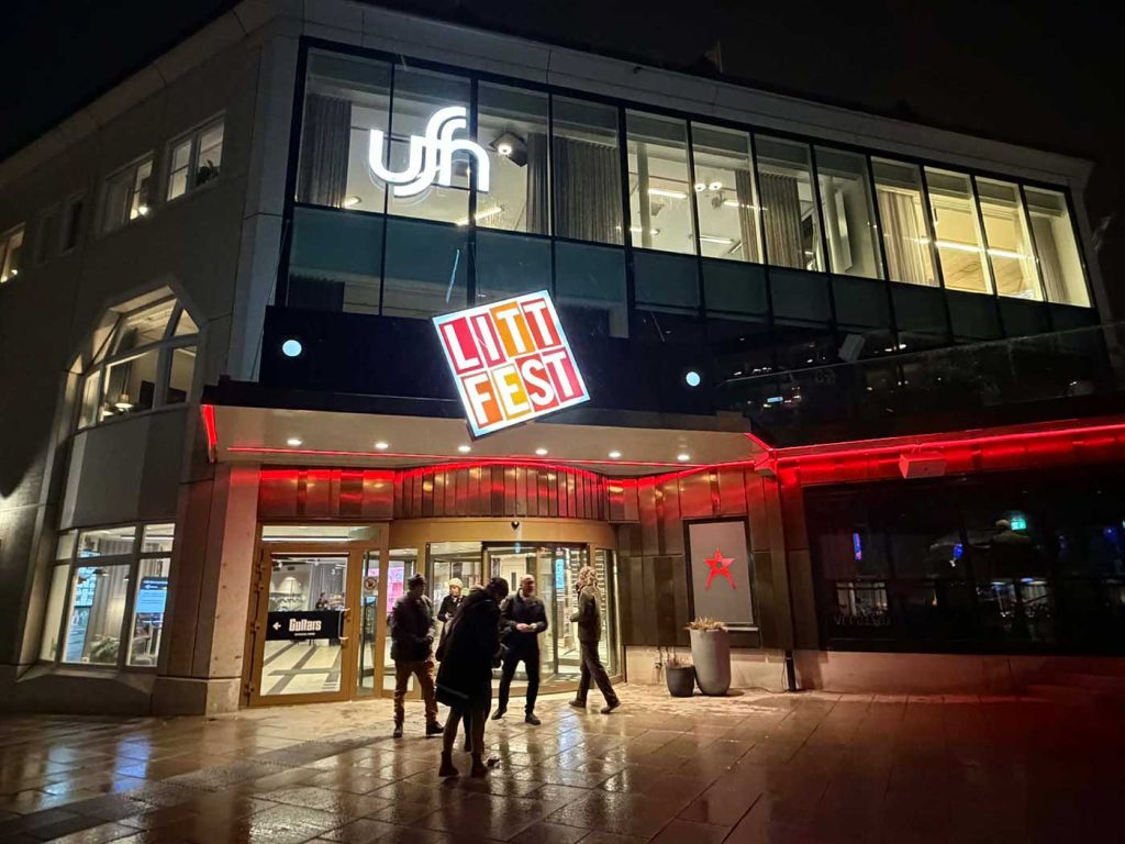 En bild av entrén till Folkets Hus Umeå. Det är kväll och mörkt. Genom fönster och glasentrén syns ljus. Ovanför glasentrén hänger en skylt med ordet Littfest. Några människor rör sig framför entrédörren.
