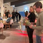 Bild inifrån en restaurang. Närmast kameran sker prisutdelning och här står en man med en blomsterbukett i handen, och en kvinna som pratar i mikrofon. Runt om i lokalen står och sitter människor som riktar uppmärksamheten mot prisutdelningen.