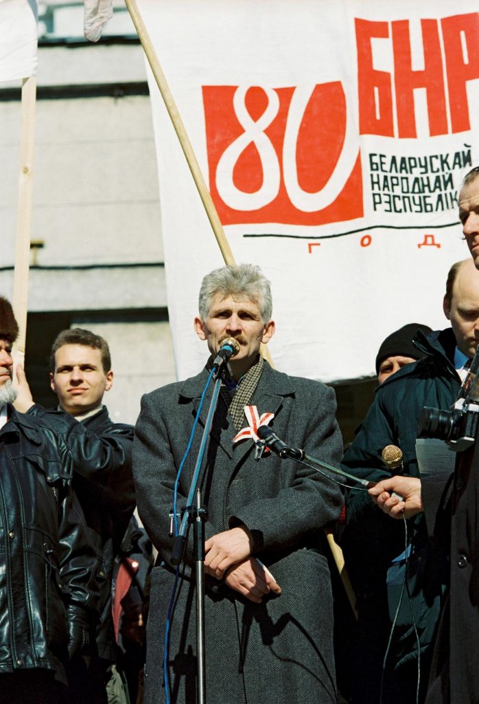 En man i grå överrock och rutig halsduk står vid en mikrofon. Bakom honom syns en affisch med ryska ord och bokstäver, och mannen är inte ensam, det finns flera män intill honom.