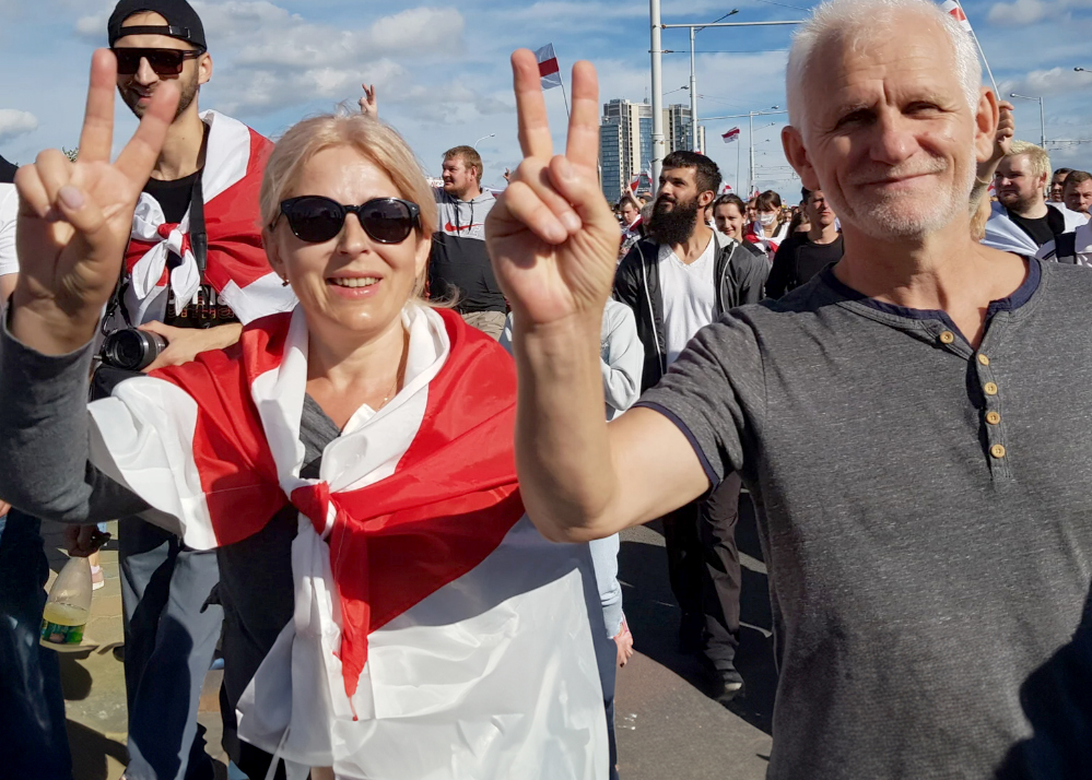 En man och en kvinna gör V-tecken med fingarna framför kameran. Dom ser ut att gå i vad som kan vara ett demonstrationståg, det ser ut att vara många människor men inga plakat syns. Dom ser glada ut, kvinnan bär mörka solglasögon. Långt bakom dem syns ett höghus. Himlen är blå med lite vita moln på. 