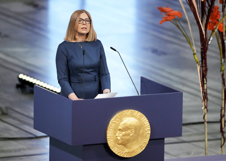 En ljushårig kvinna med glasögon står vid en talarstol och pratar i mikrofon. Talarstolen är blå och pryds av en guldfärgad plakett med Alfred Nobel. 