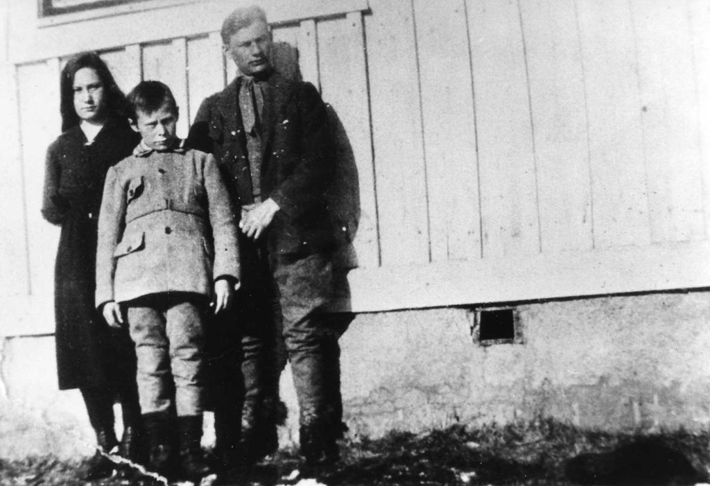 Ett gammalt svartvitt foto. En ung kvinna, en ung man och en pojke står intill en husvägg. De tittar mot fotografen och ser sammanbitna och allvarliga ut. 